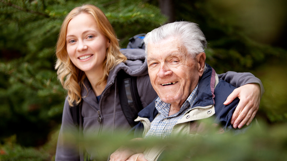 En ung kvinna håller armen om en äldre man i naturmiljö och båda ler och tittar in i kameran.